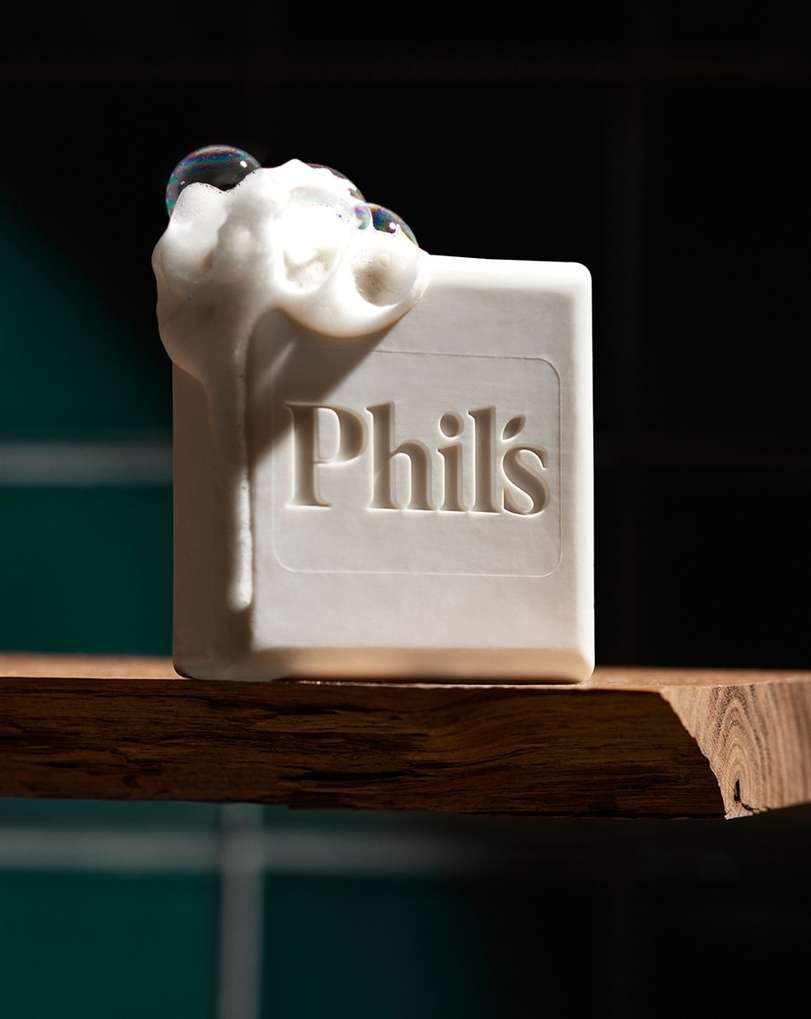 Close-up of a white bar of soap with 'Phil's' embossed on it, covered in white foam and bubbles, sitting on a wooden shelf.