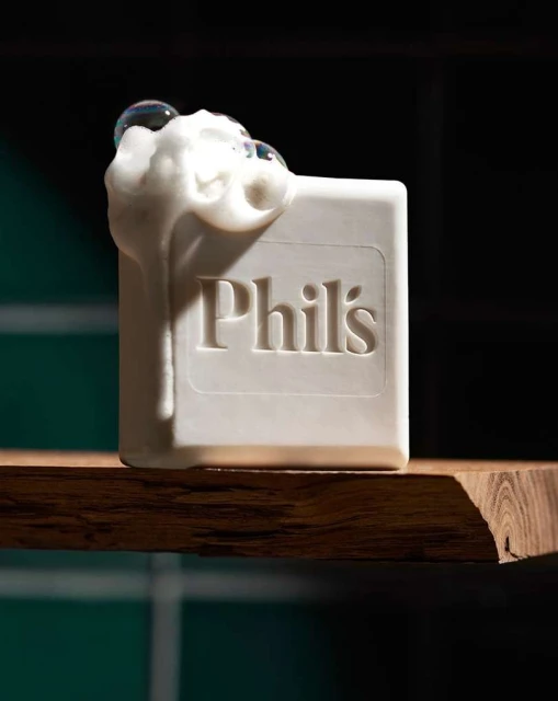 Close-up of a white bar of soap with 'Phil's' embossed on it, covered in white foam and bubbles, sitting on a wooden shelf.