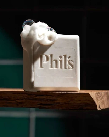 Close-up of a white bar of soap with 'Phil's' embossed on it, covered in white foam and bubbles, sitting on a wooden shelf.