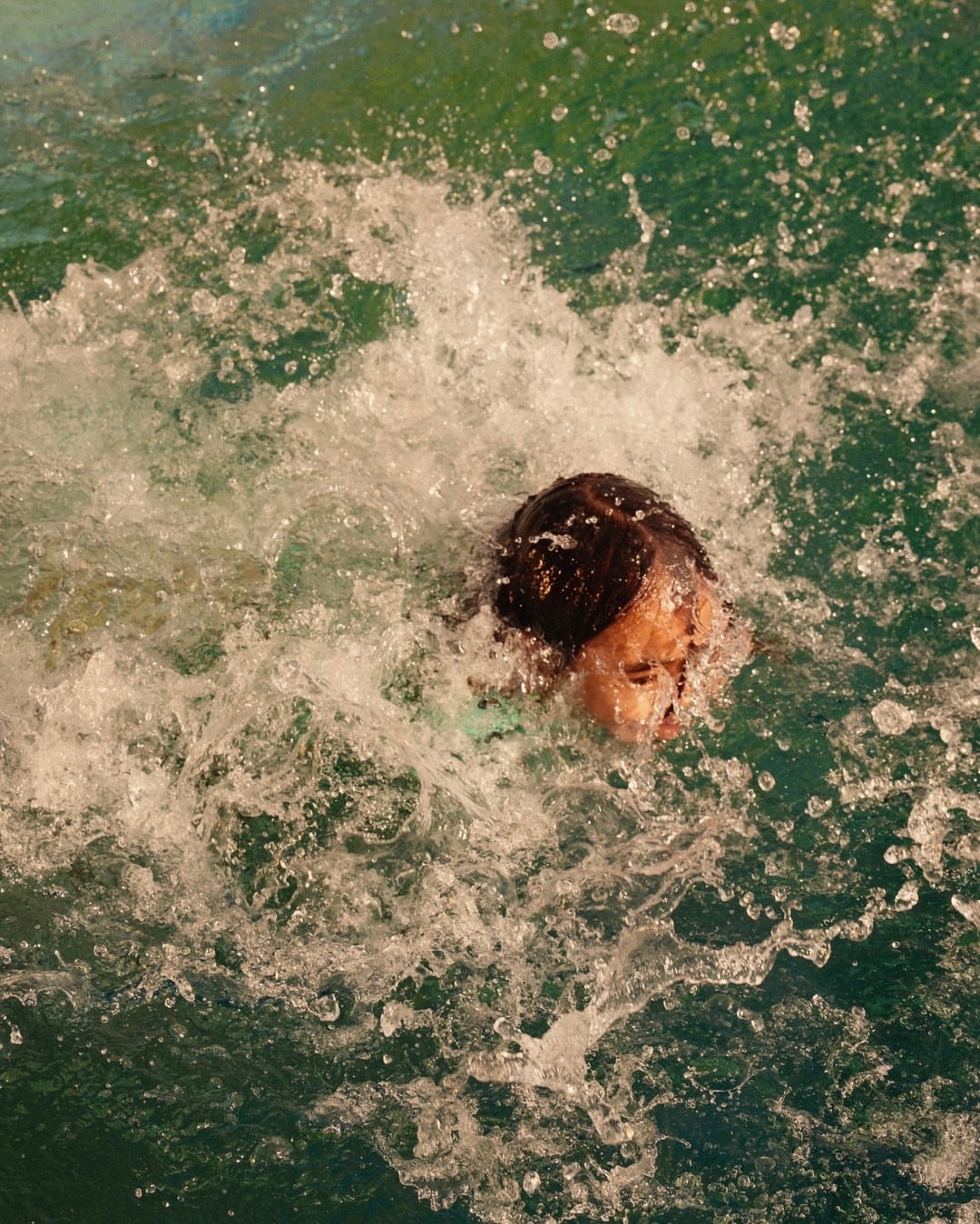 Person submerged in splashing pool water with visible face, dark hair, and turquoise pool water creating dynamic movement.