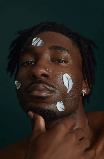 Close-up studio shot of a Black man with dreadlocks and skincare product on his face, dark green backdrop, beauty and skincare concept.