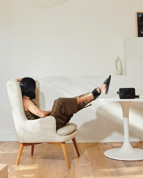 Woman relaxing in a chair with feet on table, wearing green pants and black sandals. Interior features include white walls and minimalist decor.