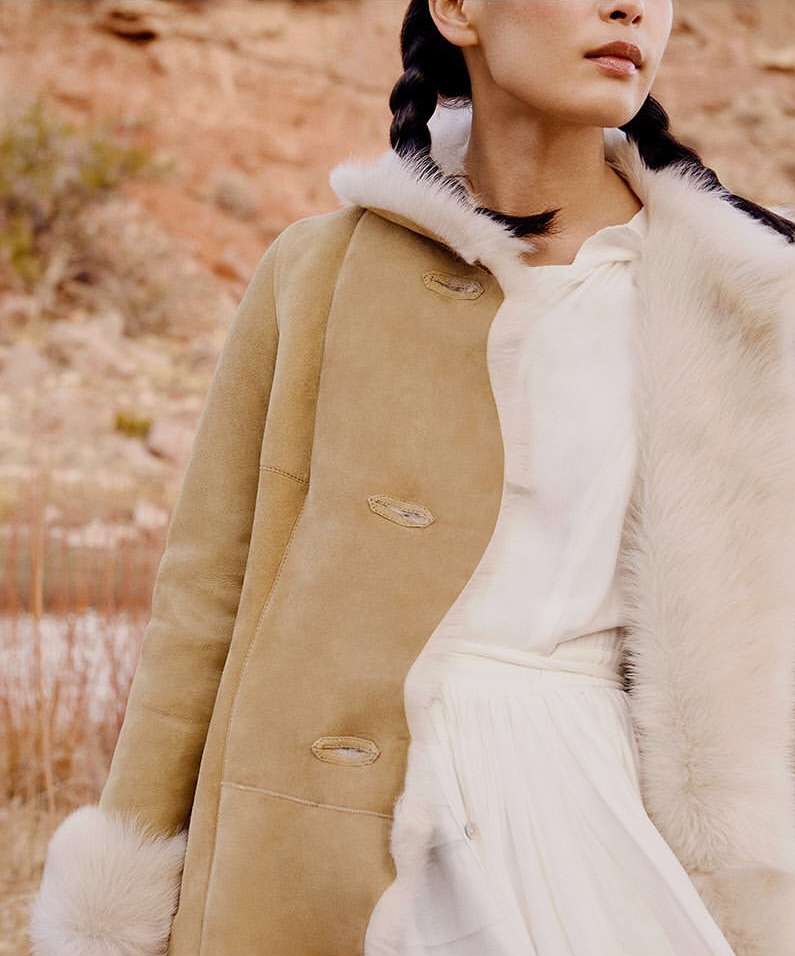 Woman in fashionable tan shearling coat with fur trim posing in a landscape with red rocks, styled hair, and neutral color palette.