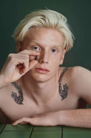 Young adult with fair skin and white-blonde hair, freckles, and dragon tattoos holding a dark oil capsule near their face.