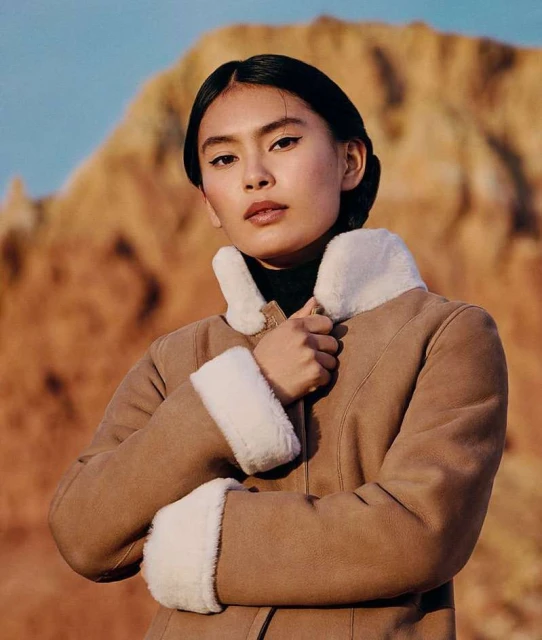 Portrait of a young Asian woman in a brown shearling coat posing outdoors against a soft background of blue sky and orange hills.