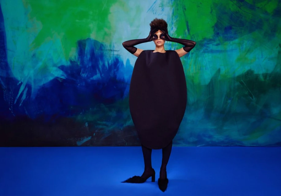 Fashion-forward photo of a woman in a black dress and gloves obscuring her eyes, set against a vibrant blue and green backdrop.