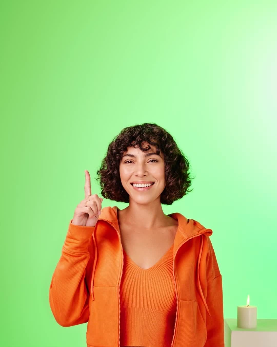 Smiling woman in orange jacket pointing upwards against a green background, with a candle visible in the lower right corner. 