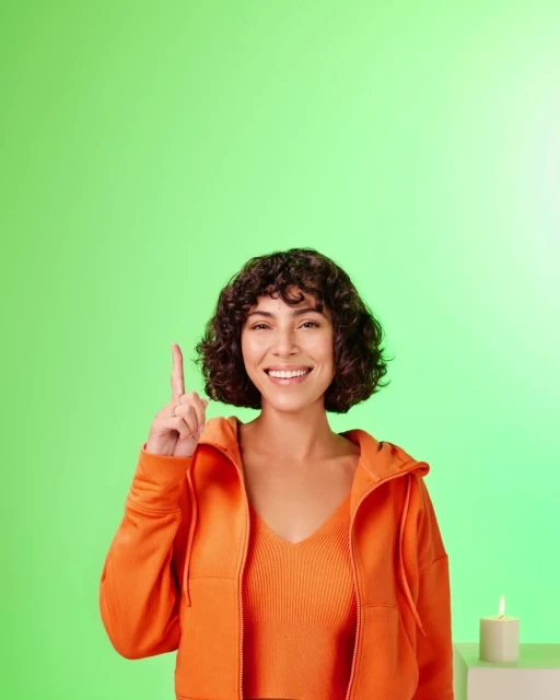Smiling woman in orange jacket pointing upwards against a green background, with a candle visible in the lower right corner. 