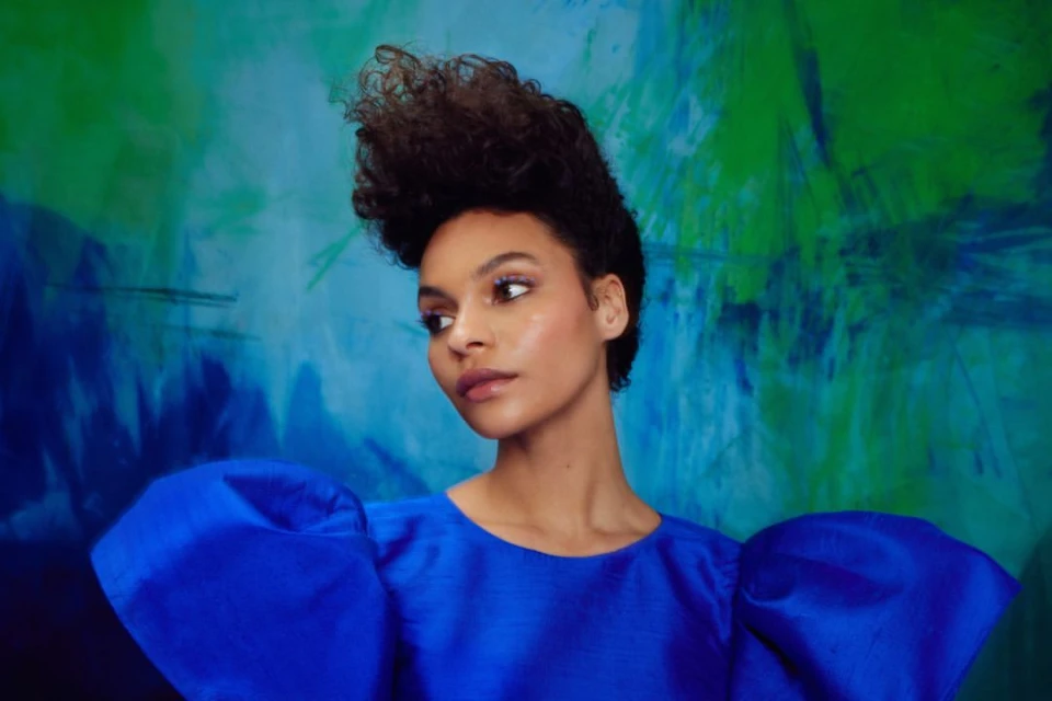 Close-up portrait of a woman with a natural hairstyle, wearing a vibrant blue dress, set against a painterly blue and green backdrop.