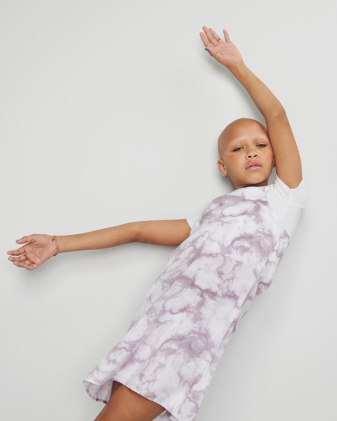 A woman in a tie-dye dress leans against a gray wall with arms extended. The dress is white with light purple tie-dye patterns.