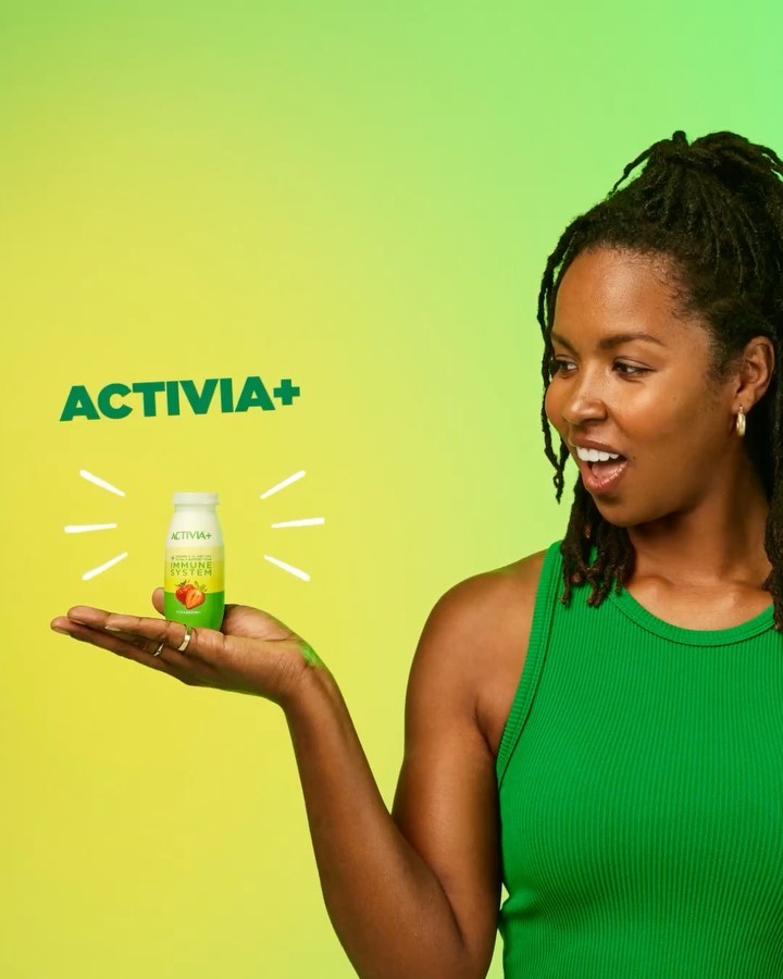 Woman with an open mouth and wearing a green shirt holds an Activia+ probiotic drink against a green and yellow gradient background.
