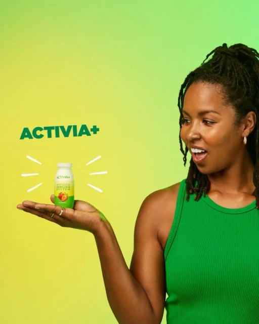 Woman with an open mouth and wearing a green shirt holds an Activia+ probiotic drink against a green and yellow gradient background.