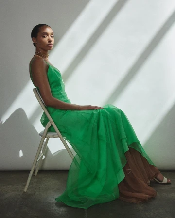 Model seated on a white folding chair wearing a vibrant green slip dress against a wall with diagonal sunlight shadows