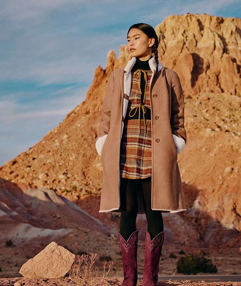 Asian woman stands in the desert wearing a beige coat over a plaid dress with burgundy cowboy boots against a desert landscape backdrop.
