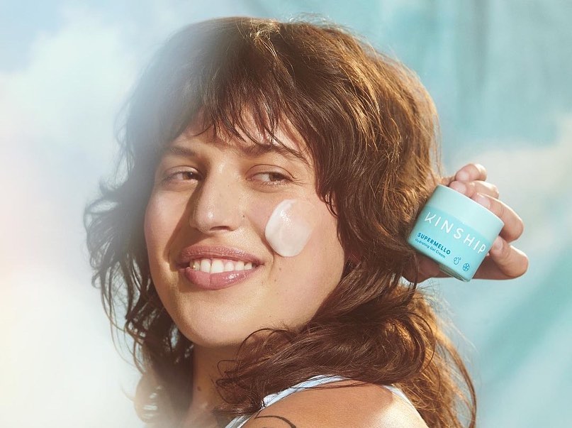 Woman with skincare product on her face and holding Kinship Supermello Hydrating Gel Cream in an aqua-colored container, against a light blue background.