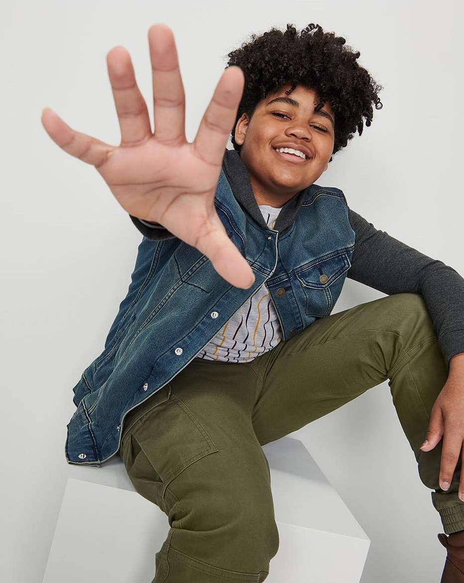Smiling young person in a denim vest and olive-green pants extends their hand toward the viewer while sitting on a white cube.