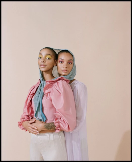 Two women in fashion headscarves and pastel outfits embrace in front of a beige background, creating a stylish studio portrait.