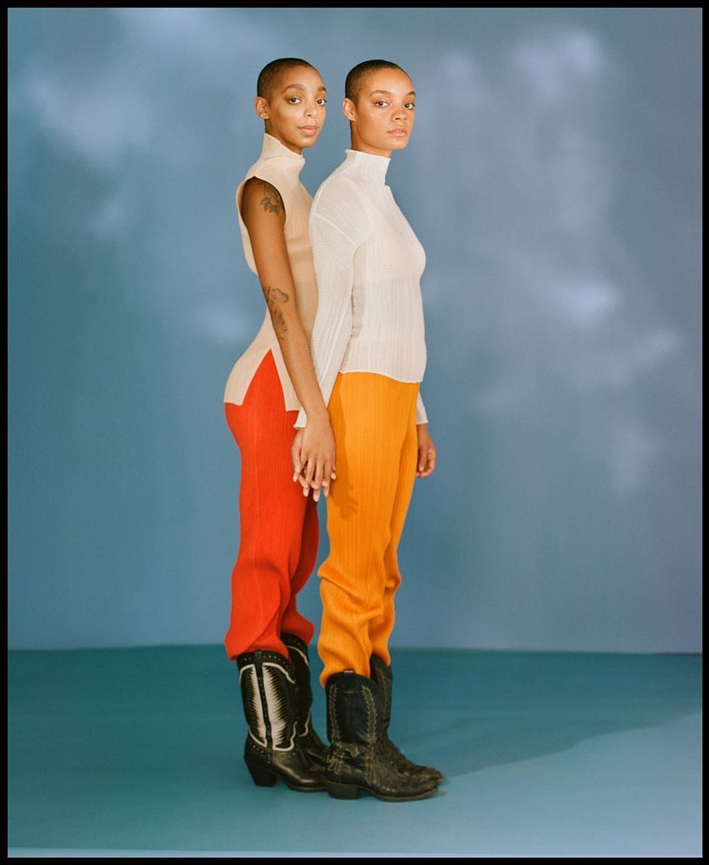 Two short-haired women in turtlenecks and colorful pants stand close against a blue backdrop, holding hands and wearing cowboy boots.