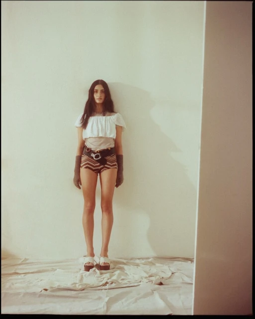 Full-length shot of a woman in a white crop top and patterned shorts posing against a white wall in a photo studio.