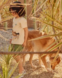 Young child in cream t-shirt and green shorts standing outdoors beside a large golden dog