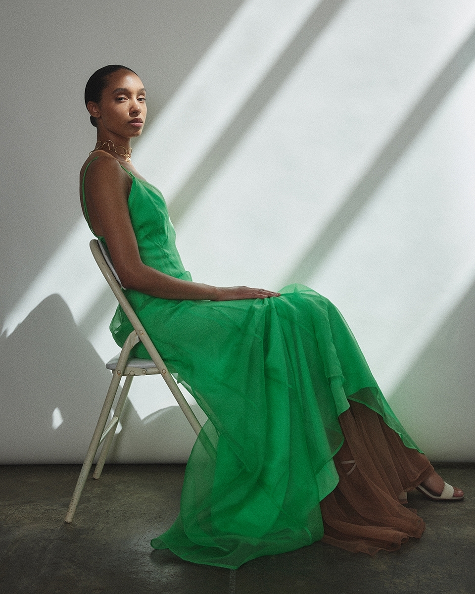 Model seated on a white folding chair wearing a vibrant green slip dress against a wall with diagonal sunlight shadows