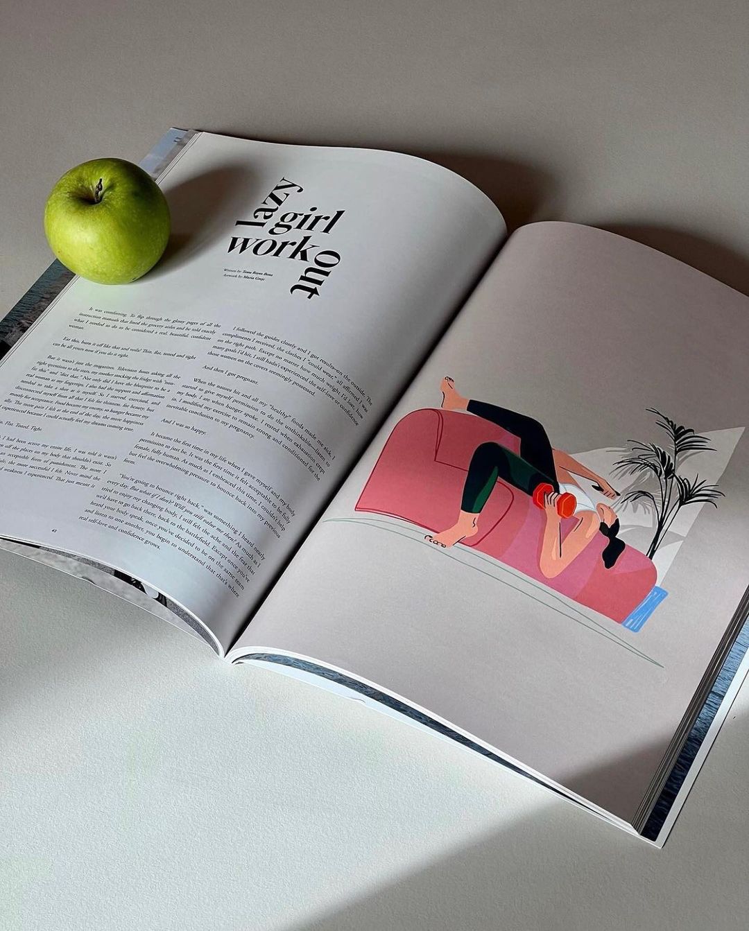 Open magazine displays an illustration of a woman on a pink couch, with an apple on the adjacent page, offering a lifestyle scene.
