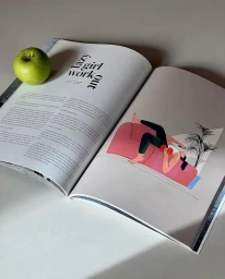 An open magazine page featuring editorial illustration by Maria Grejc, resting on a plain surface beside a green apple.