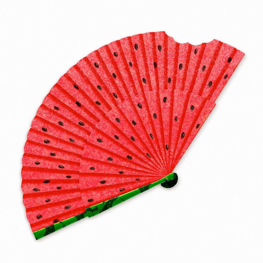 Watermelon-themed paper fan on a white background. The fan is red with black seeds and a green handle. The image is well-lit.