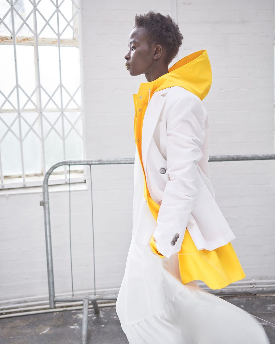 Fashion shot of an adult female walking, wearing a white blazer and a yellow hoodie, with natural light, in an indoor location.