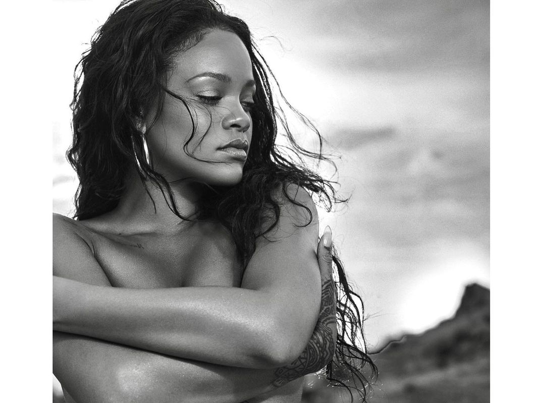 Black and white portrait of Rihanna with closed eyes, arms crossed, wearing hoop earrings, set against a blurred mountain background.