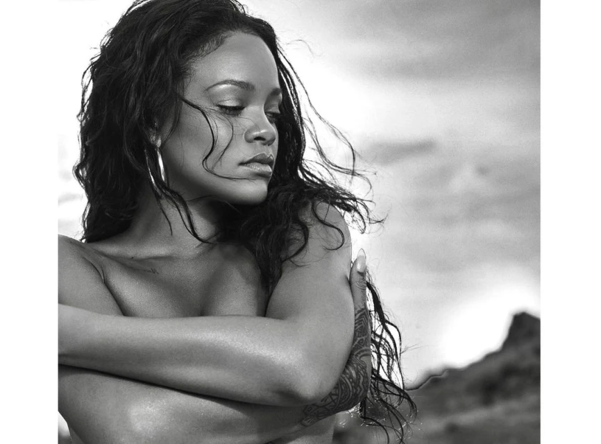Black and white portrait of Rihanna with closed eyes, arms crossed, wearing hoop earrings, set against a blurred mountain background.