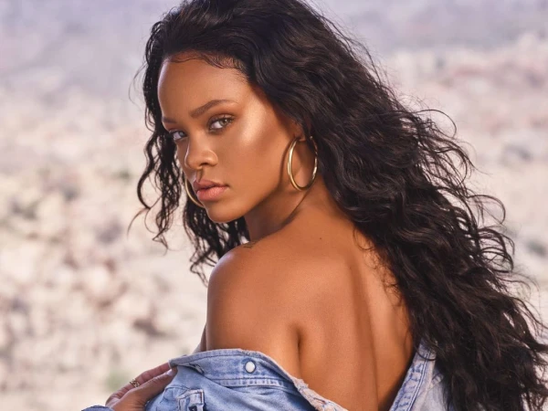 Rihanna wearing a denim jacket and large gold hoop earrings, looking over her shoulder against an outdoor mountainous backdrop.