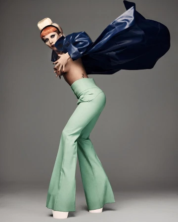 Lady Gaga in high-waisted green trousers and a billowing blue cape against a plain grey studio background.