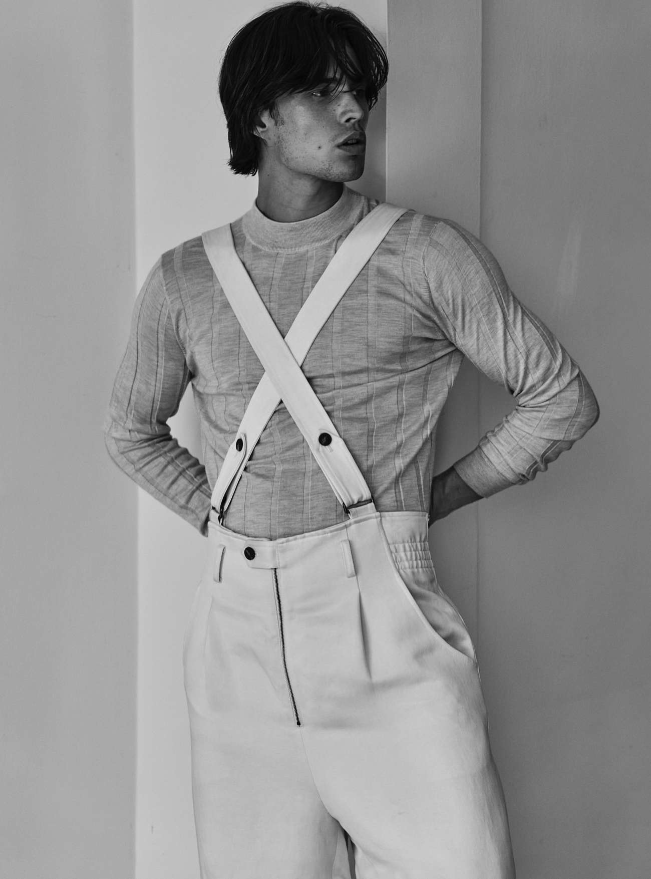 Black and white portrait of a young adult man wearing a long-sleeved shirt and suspenders, standing against a neutral-toned wall.