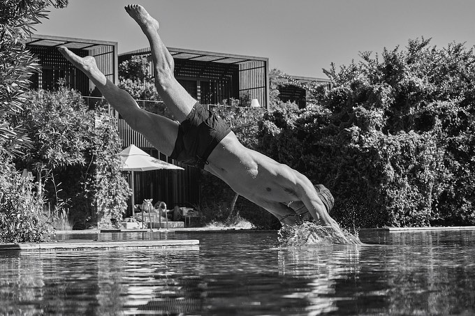 Monochrome photo of athletic adult male diving headfirst into a pool, with lush greenery and architecture in the background, sunny day.