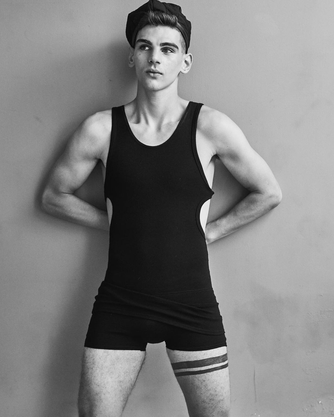 Black and white portrait of a fit, young adult male wearing a swim brief, tank top, and newsboy cap against a neutral wall backdrop.