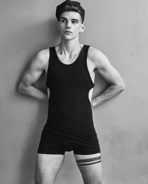 Black and white portrait of a fit, young adult male wearing a swim brief, tank top, and newsboy cap against a neutral wall backdrop.