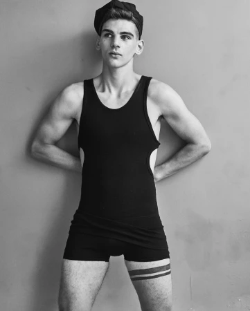 Black and white portrait of a fit, young adult male wearing a swim brief, tank top, and newsboy cap against a neutral wall backdrop.
