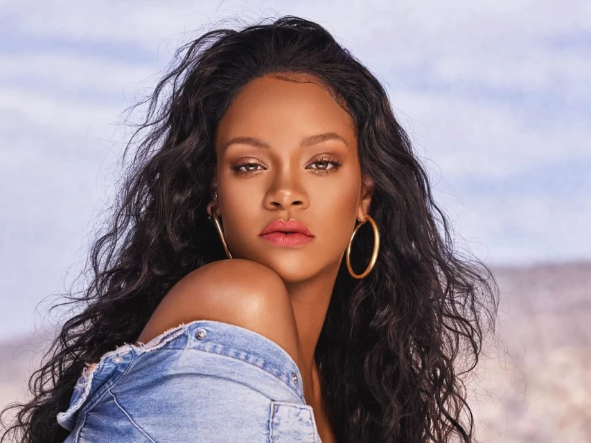 Close-up of a model with long, wavy black hair, hoop earrings, and a denim jacket against a blurred outdoor backdrop with sky.