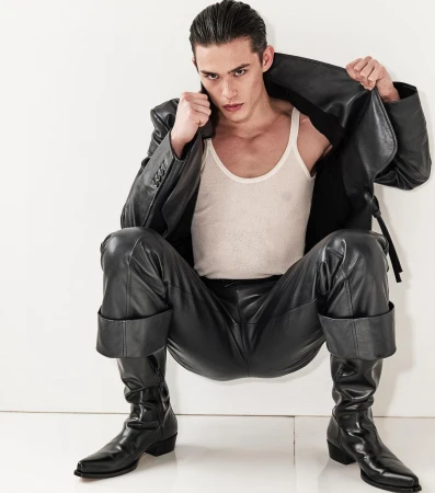 Young male model crouching against a white background wearing black leather trousers, boots, a jacket, and a white tank top.