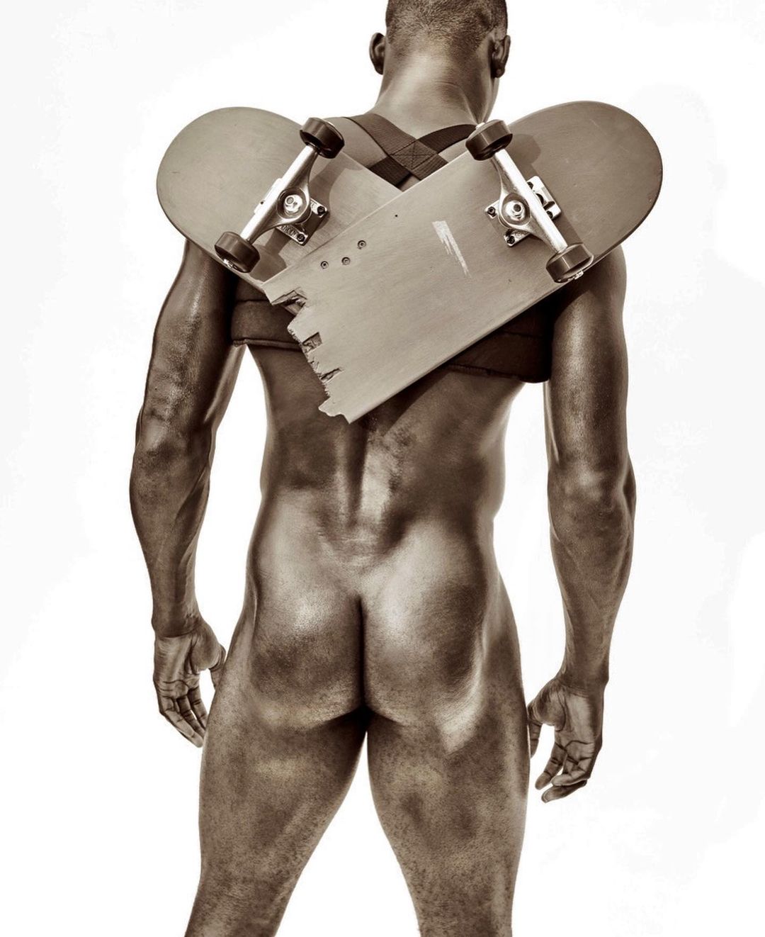 An undressed Black adult male faces away in a studio, with a broken skateboard strapped to their back by black straps over the shoulders.
