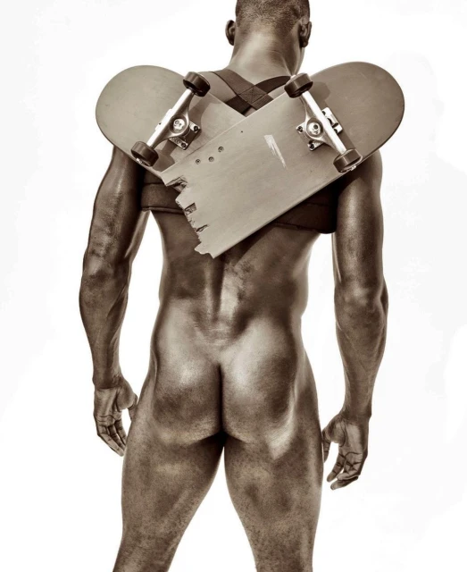 An undressed Black adult male faces away in a studio, with a broken skateboard strapped to their back by black straps over the shoulders.