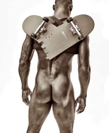 An undressed Black adult male faces away in a studio, with a broken skateboard strapped to their back by black straps over the shoulders.