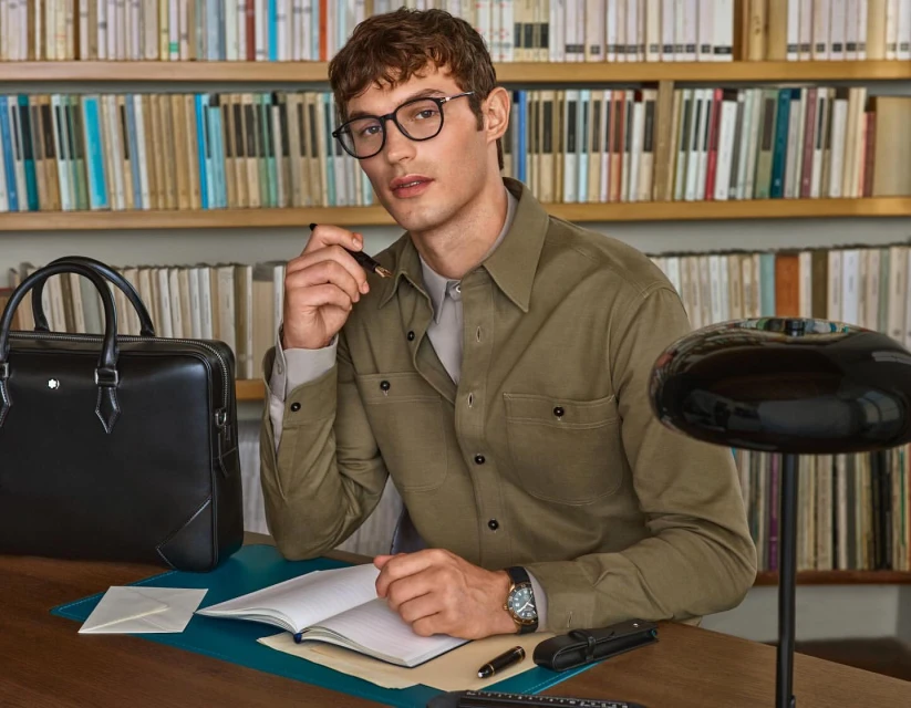Man sits at a desk in front of a bookshelf holding a pen, wearing a button-down shirt with glasses, and a black leather briefcase.
