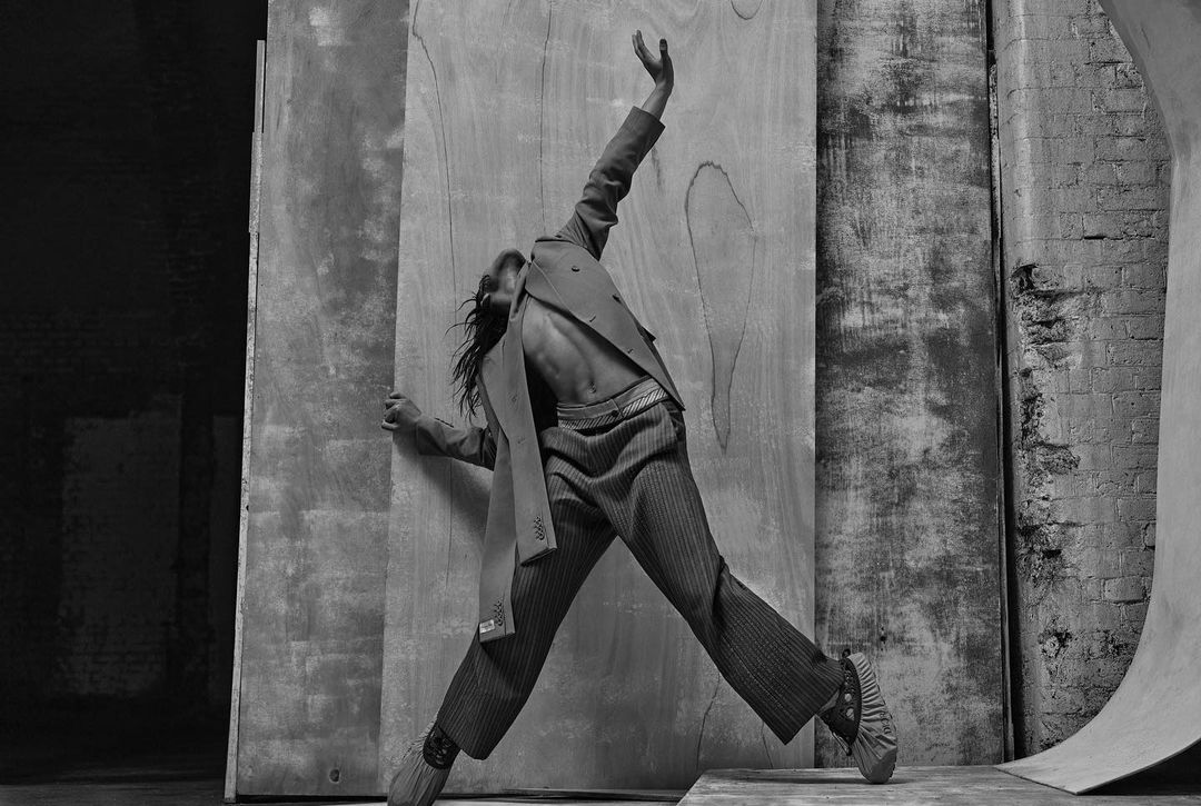 Black and white fashion shot: a person in a blazer and striped pants strikes a dynamic pose against a textured backdrop with dramatic lighting.