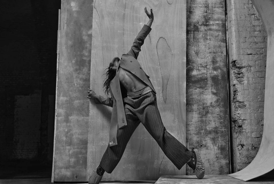 Black and white fashion shot: a person in a blazer and striped pants strikes a dynamic pose against a textured backdrop with dramatic lighting.