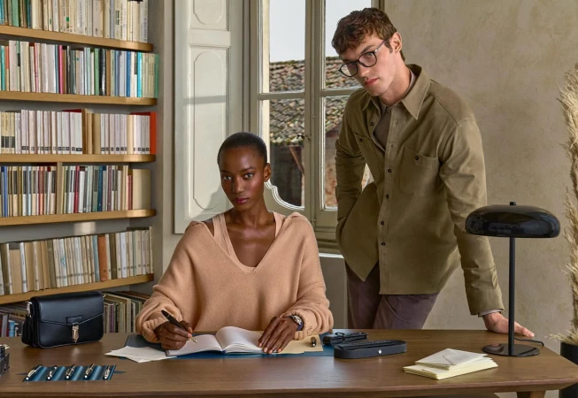Indoor shot of an African woman writing in notebook at a wooden desk with fountain pens, next to a caucasian man standing near a window.