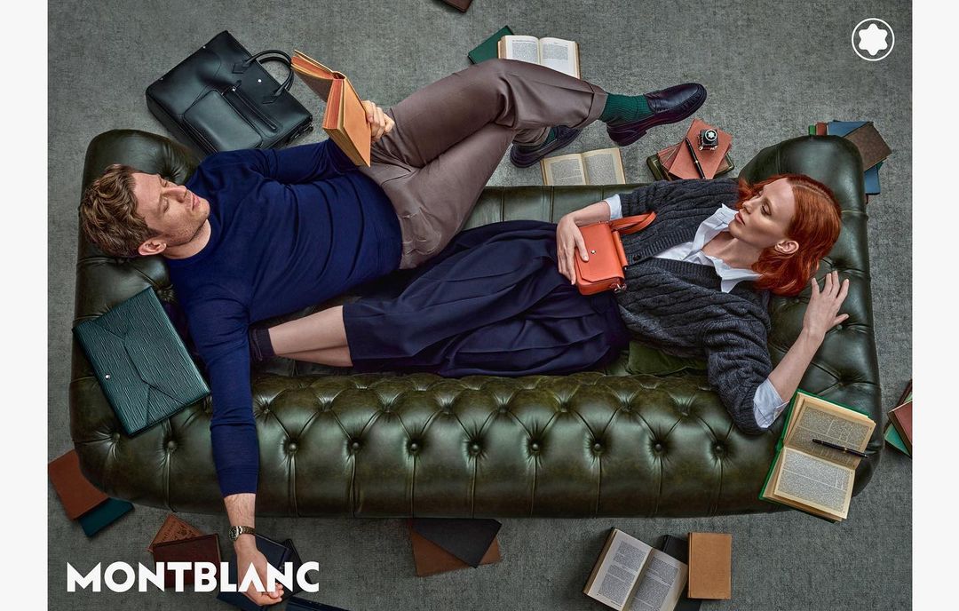 Man and woman lounge on an olive-green sofa, surrounded by leather accessories and open books in a fashion-focused editorial shot.