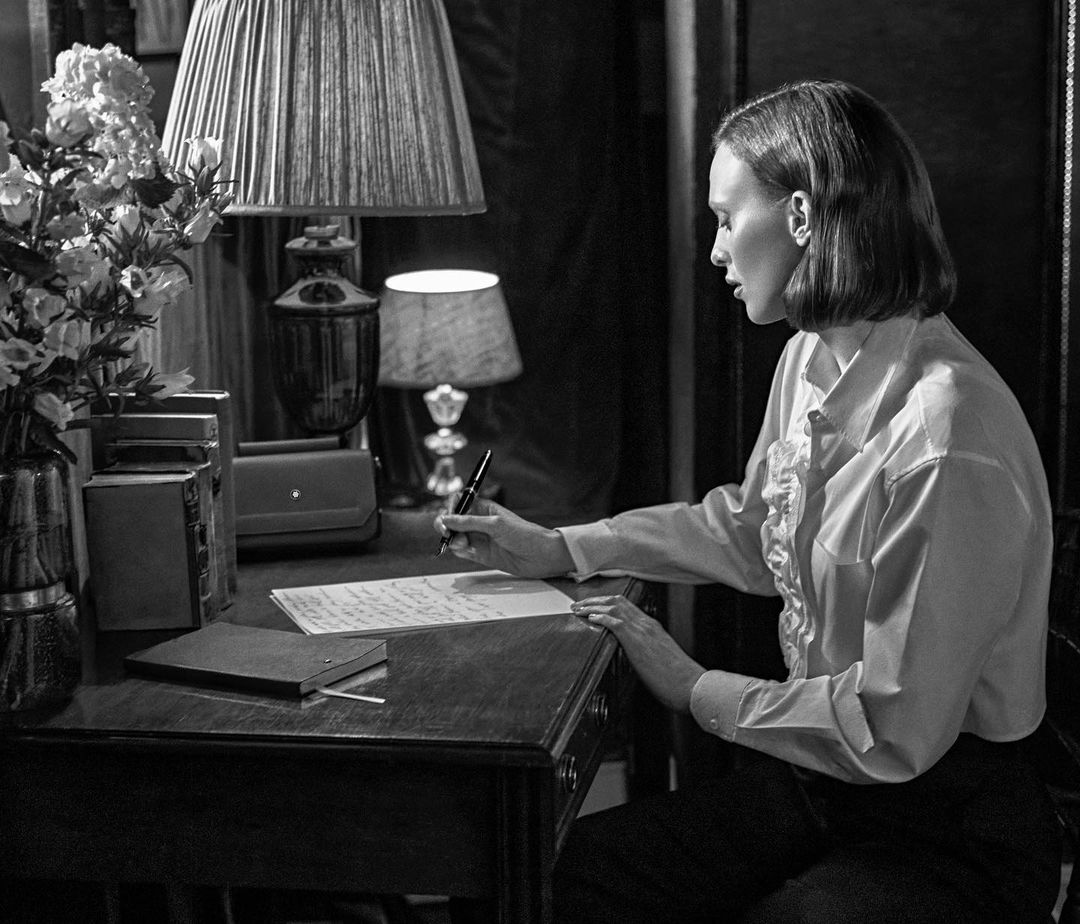 Monochrome portrait of a woman in a ruffled blouse writing at a wooden desk with flowers, books, and lamps; vintage style.