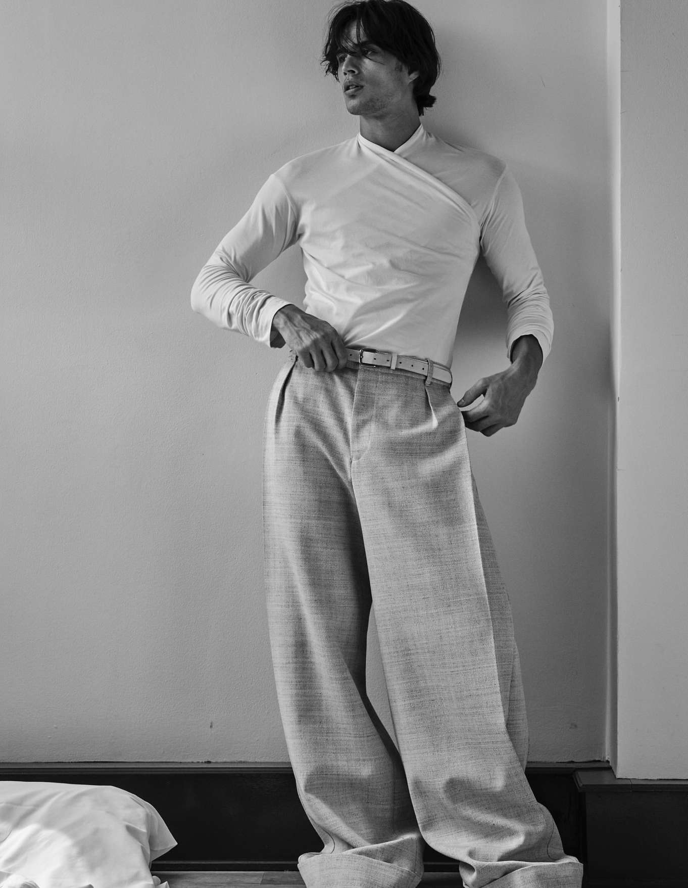 Monochrome portrait of a male model in a fitted long-sleeve top and wide-leg trousers leaning against a wall.